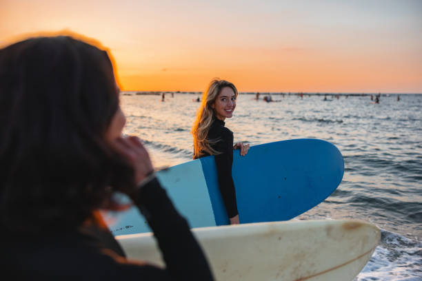 Attractive young person caucasian blonde female surfer looking back while walking towards the sea. She is accompanied by her best friend. They are going for an evening surfing session during sunset. The girls are wearing black wetsuits and are carrying their surfboards.